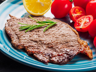 Close up view of fried steak served with fried potatoe sherry tomatoes in blue plate on black concrete background