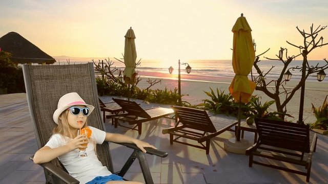 Young caucasian girl sitting on armchair and drinking orange juice cocktail through straw. Sea coast horizon at backgrounds. Girl drinking orange juice sitting on armchair. Vacation resort.