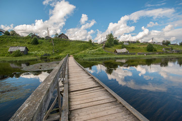 north Russian village Isady. Summer day, Emca river, old cottages on the shore, old wooden bridge and clouds reflections.