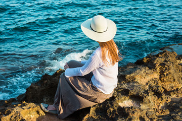 Young beautiful woman in boho clothes long hair sunhat sitting on rocks at the shore looking at the sea, meditation, tranquility, harmony, serenity