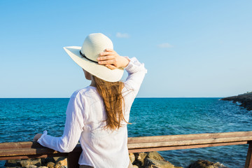 Young beautiful slender woman with long hair holding sunhat from wind in boho style clothes at the shore looking and the sea clear blue sky, rocks