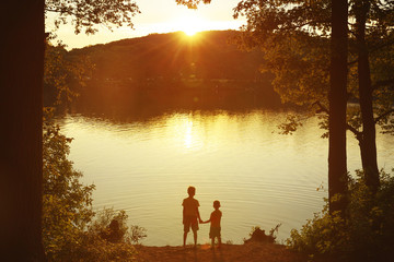 Children at the water watching the setting sun. Two boys holding hands standing on the shore. Back...