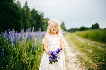 Fototapeta na wymiar Lost, lonely and beautiful baby kid little blonde girl with offended pathetic poor tear-stained face standing on field with flowers road at nature outdoor in white dress and holding bouquet of lupines