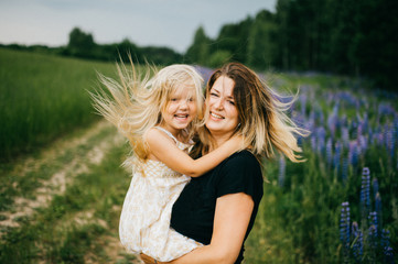 Fototapeta premium Young mother with long hair holding little daughter princess at hands. Playing with child. Pure, happy, kind, expressive emotional faces. Family at nature. Artistic kid. Laughing, smiling, embracing.