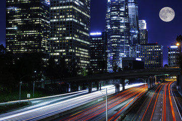 Fototapeta na wymiar cars traveling on freeway at night among Los Angeles downtown high rises with full moon in sky