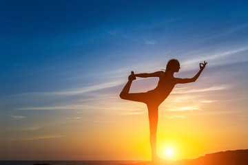 Silhouette of young woman doing fitness exercises on the sea beach during amazing sunset. Yoga and healthy life style.