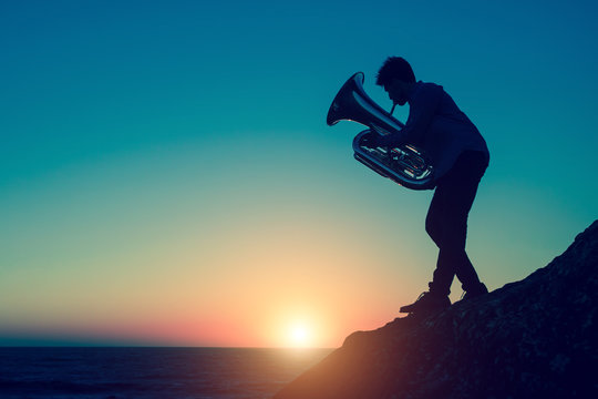 Silhouette of musician playing the tuba on rocky sea coast during amazing sunset. Trumpet.