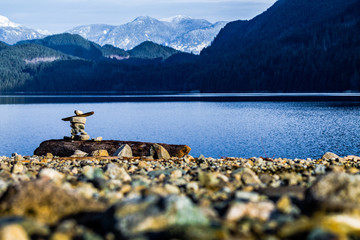 inukshuk on rocky shore with mountains and lake - obrazy, fototapety, plakaty