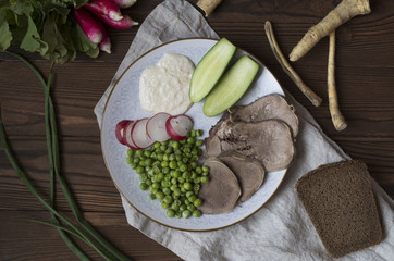 beef tongue with radish, cucumber, green pea and sauce on a plate with fresh radish, bread, onion on a brown wooden table, top view