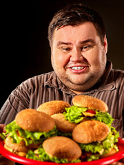 Fat man eating fast food hamberger and carries treat for friends on tray. Breakfast for overweight person. Poor food leads to obesity. Person regularly overeats concept on black background.