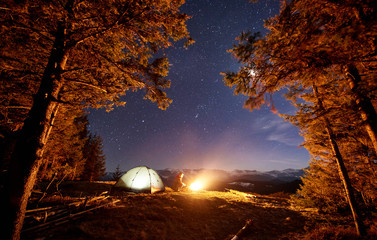 Male hiker have a rest in his camp near the forest at night. Man sitting near campfire and tent under beautiful night sky full of stars and the moon and enjoying night scene