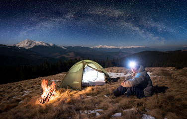Male tourist have a rest in his camp at night. Man with lighting headlamp sitting near campfire and tent, looking to the camera under beautiful sky full of stars and milky way