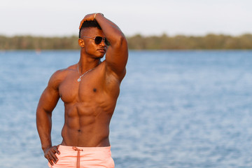 A young athletic beautiful guy with a muscular body. A black sports man is practicing on the beach at sunset. Muscular guy, sportsman, fitness model. The boy in the sunglasses on the beach.