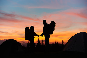 Silhouettes of a man and woman holding hands standing on top of the mountain while hiking together with their backpacks enjoying beautiful sunset with fiery colorful sky love relationships nature.