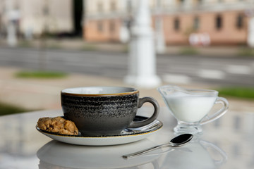 A grey cup of black coffee and a cookie on a saucer, a spoon, a milk jug, on a marble table. City cafe terrace. Minsk, Belarus. Selective focus.