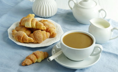 A white cup of coffee with milk and homemade sugar cookies bagels on blue linen tablecloth and white wooden table