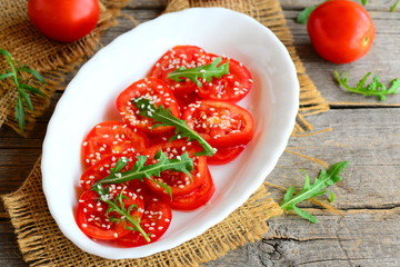 Easy tomatoes, rucola and sesame seeds salad. Bright veggie salad on a white plate and burlap textile. Fresh red tomatoes and green rucola on an old wooden table
