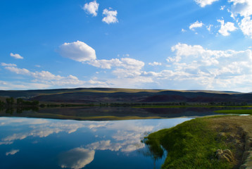 View of calm lake, hills, green grass and blue sky in Altai mountains. White clouds reflected in water. Chuya steppe, Altay Republic, Siberia, Russia.