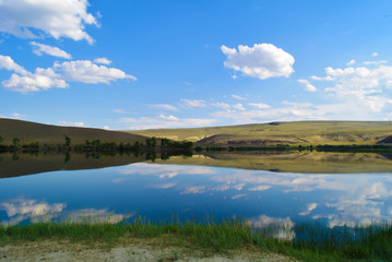 View of quiet lake, hills, green grass and blue sky in Altai mountains. White clouds reflected in water. Chuya prairie, Altay Republic, Siberia, Russia.