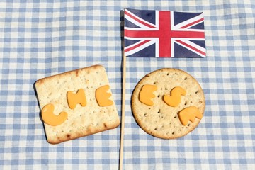 The word cheese cut out from Red Leicester cheese on crackers with a Union Jack flag on a blue gingham tablecloth