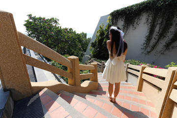 back view of asian woman who wear white dress with little wing stand on way down of stair case
