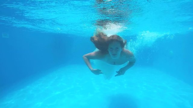 Young girl in white wedding dress swims under water in swimming pool. Underwater shot of teen girl with long blonde hair diving in slow motion.
