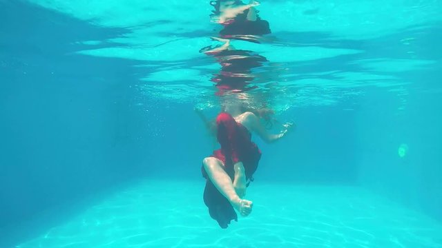 Young girl in long red dress swims under water in swimming pool. Underwater shot of long-haired teen girl diving in blue water of the pool. Slow motion.