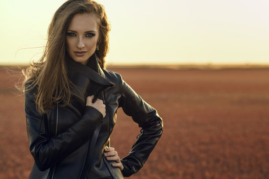 Young beautiful glam model with the wind in her long hair wearing black stylish leather jacket standing in the deserted field at sunset wrapping her oversized collar tighter. Copy-space
