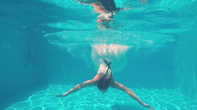 Young girl with braided pigtails in white wedding dress swims under water in swimming pool. Underwater shot of diving girl in blue water of the pool. Slow motion.