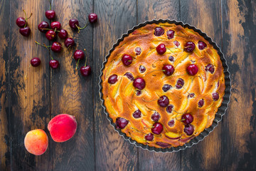 Homemade fruit peach and sweet cherry cake in a black baking dish on the rustic wooden table, top view.