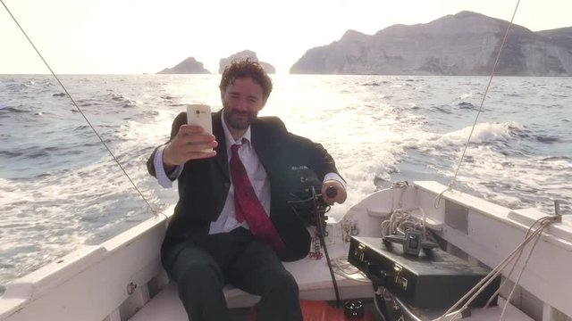Man in suit with a briefcase texting on his smartphone while driving a boat in the middle of the ocean