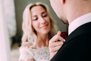 Bride fixes red bow tie on groom's neck while they stand near the window