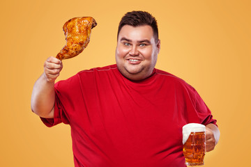 Not sporty fat man at oktoberfest, drinking beer and eating chicken leg on yellow background.