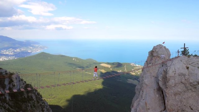 Brave Woman Walking on a Scary Suspension Bridge. Popular Russian Touristic Mountains Ai-Petri, Crimea. Black Sea Coast and Yalta City on Backgroung. HD.