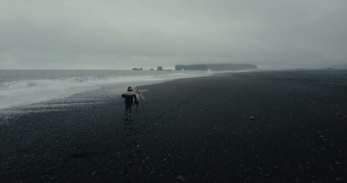 Copter flies after young couple running on the shore of the sea in icelandic sweater. Black volcanic beach in Iceland.