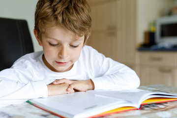 Little blonde school kid boy with glasses reading a book at home