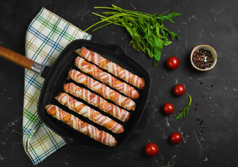 Preparation of raw sausages wrapped spirally in bacon on a cast-iron frying pan. Ingredients for sausages in bacon cherry tomatoes, rucola salad. Black concrete background. Top view, flat lay.