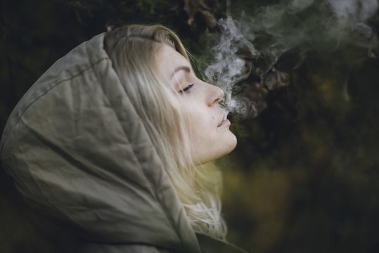 Young blonde woman with a hood on blowing smoke out her mouth