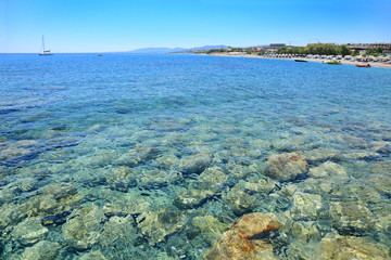 Kolorowe kamienie w przeźroczystej wodzie morza Śródziemnego, plaża, parasole, hotele.