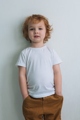 Little cute redhead curly boy in a white T-shirt, on a white background. Keeps his hands in his pockets