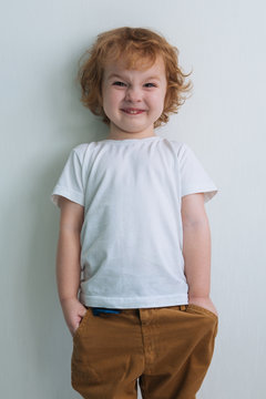 Little cute redhead curly boy in a white T-shirt, on a white background. Keeps his hands in his pockets