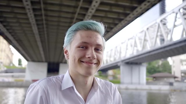 portrait of joyful happy teenager with blue hair. he has pimples on the face and crooked teeth in the mouth. walking around the city