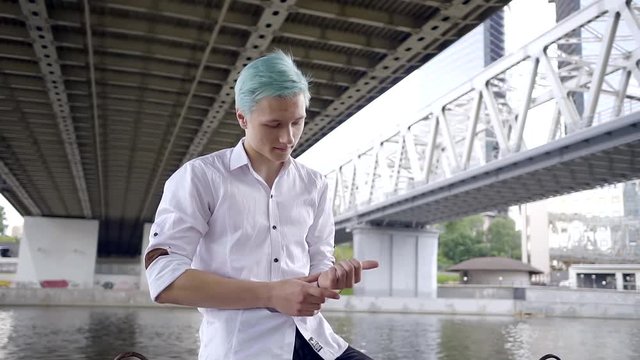 young stylish boy gay with blue hair in a white shirt sits under a highway bridge across the river