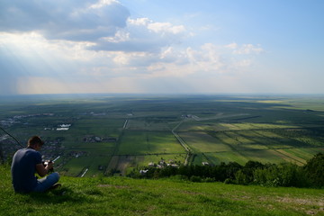 A young man siting on a hill and photographing amazing view of the plain