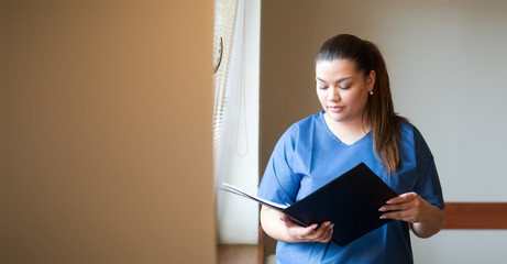 Young hispanic nurse reading a clinical report in a hospital hallway, wearing blue uniform.