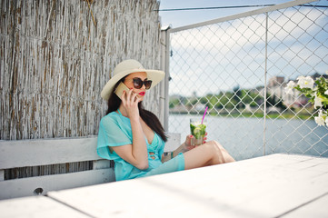 Sweet young lady wearing hat and sunglasses with a cocktail using her smartphone while sitting by the lake.