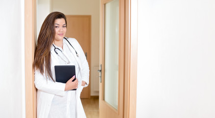 Female young hispanic doctor holding a digital tablet, standing in a hospital hallway, smiling and looking into camera with lots of white copy space