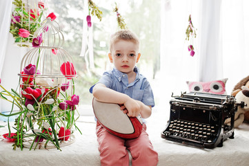 Portrait of a stylish and hndsome little boy sitting on the windowsill with flowers and printing machine.