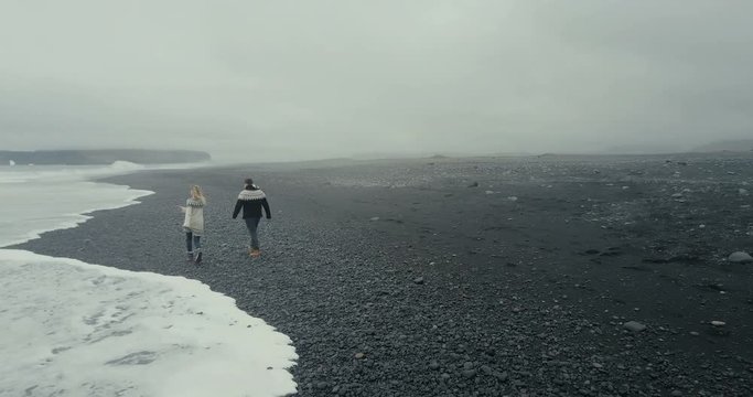 Aerial view of young couple walking on the black volcanic beach in Iceland. Man and woman in lopapeysa throw rock on sea