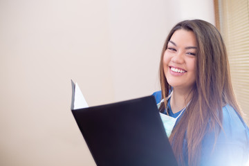 Close-up of smiling confident medical female doctor standing in a hospital hallway next to a window, wearing a blue uniform and stethoscope, holding a folder, looking into camera